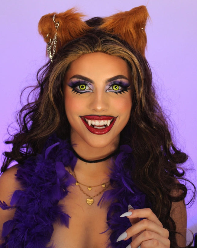 Portrait of a woman in a glamorous werewolf-inspired costume. She has bright green cat-like contact lenses, sharp fangs, and bold makeup featuring dramatic purple eyeshadow and dark red lips. Her long wavy brown hair is complemented by furry wolf ears adorned with small chains. She wears layered gold necklaces, a black choker, and a purple feather boa. Her expression is playful with a wide, fang-bearing smile, and she poses against a soft purple background.